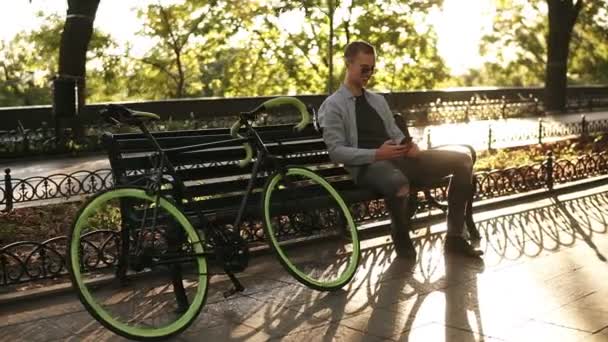 Hombre joven con camisa azul sentado en el banco y sosteniendo su teléfono móvil en las manos, mensajes de texto. Jinete tomando un descanso después de montar en bicicleta en su bicicleta de trekking en el parque. El sol brilla, árboles verdes en el — Vídeos de Stock
