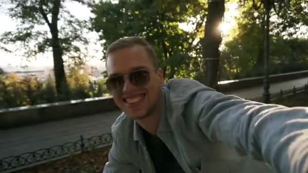 Joven alegre en bicicleta en el parque de la ciudad en un hermoso día de verano. Niño sonriente en gafas de sol disfrutando del aire libre montando y filmando su día activo con su teléfono móvil — Vídeo de stock