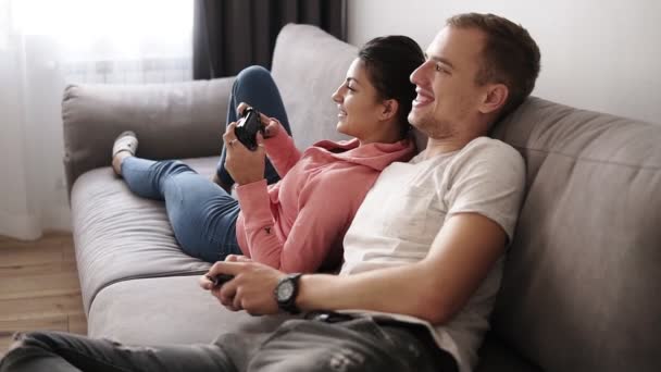 Entspanntes junges Paar, das zu Hause Videospiele spielt, während es auf dem Sofa liegt und sich amüsiert. Sie haben ein tolles Wochenende drinnen und spielen Fernsehspiel mit Steuerknüppeln. Seitenansicht — Stockvideo