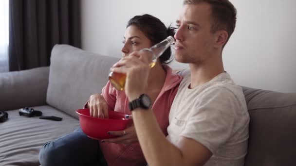 Młoda para oglądając telewizor w domu siedząc na kanapie i cieszyć się pokazuje. Mają one przekąski - jedzenie chipsów i picia piwa. Pomieszczeniu — Wideo stockowe