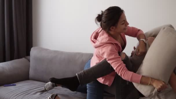 Joven pareja caucásica divirtiéndose y disfrutando de un tiempo de calidad juntos mientras están sentados en el sofá en casa. La chica está jugando pelea de almohadas con su novio y sonríe. Movimiento lento — Vídeo de stock