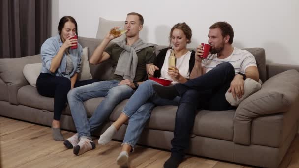 Los amigos se reúnen y se divierten en la sala de estar con el interior del loft. Compañía masculina y femenina, la chica con un tazón rojo grande con palomitas de maíz, todo el mundo bebiendo cerveza o refresco, riendo — Vídeo de stock