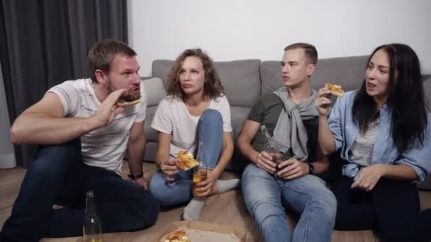 Nourriture, loisirs et friandises - quatre jeunes souriants mangeant de la pizza à la maison et buvant une bière assis sur le sol. Homme barbu racontant des histoires drôles, tout le monde riant, ayant bon — Video