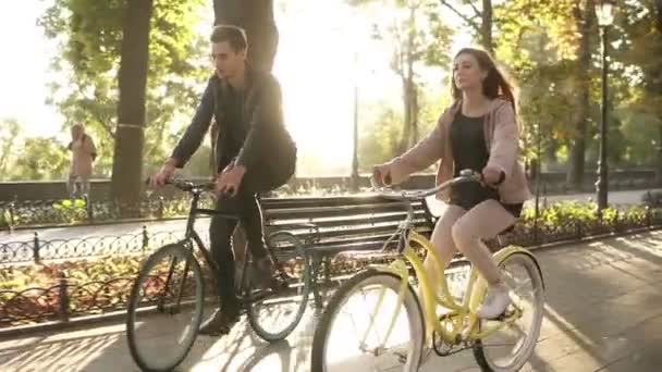 Close up of caucasian young couple or friends riding their bikes in the empty city park or boulevard in summertime. People, leisure and lifestyle concept. Green trees around, sun shines on the — 图库视频影像