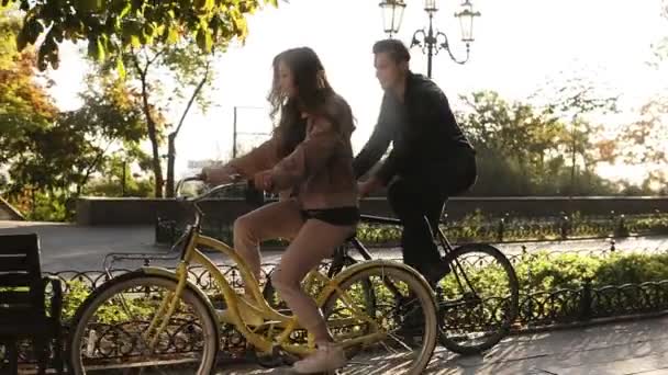 Hermosa pareja joven montando sus bicicletas en el parque vacío de la ciudad. Detente y siéntate en el banco y abrázate. Una pareja amorosa. El sol brilla en el fondo — Vídeo de stock