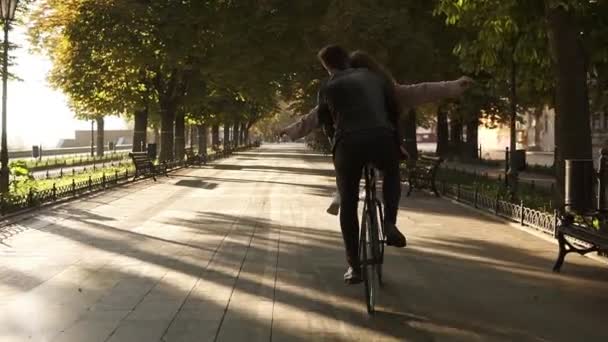 Ungt par ha kul ridning på samma cykeln i utomhus aktivitet med sun bakgrundsljuset på bakgrunden. Flicka sitter på rodret med utsträckta händer. Människor, romantik, fritid koncept. Sällsynt vy — Stockvideo