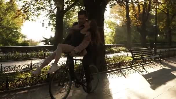 Aimer les jeunes couples ont plaisir à rouler sur le même vélo dans l'activité de plein air avec le soleil rétroéclairage sur le fond. Fille est assis sur le gouvernail et embrasser son petit ami — Video