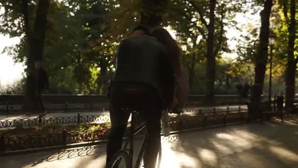 Gioioso giovane coppia in sella alla stessa bici nera con retroilluminazione solare e alberi sullo sfondo. Ragazza è seduta sul timone mentre il suo fidanzato in bicicletta. Vista rara — Video Stock