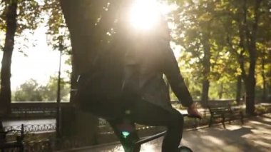 Siyah rahat kıyafetler, kulaklık arkasında siyah durumda gitar ile şehir parkta Bisiklete binmek genç bir erkekte nadir bir bakış. Yeşil ağaçların çevresinde