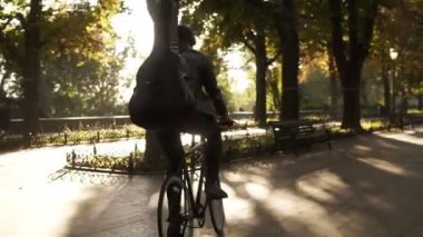 Siyah rahat kıyafetler, Bisiklete binmek tarafından döşeli Şehir Parkı gitar siyah durumda ile arkasında ve müzik kulaklık genç bir erkekte nadir bir bakış. Yeşil ağaçların çevresinde