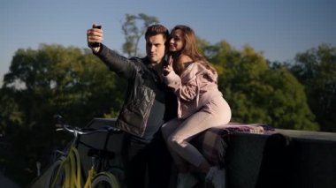 Güzel genç çift alarak selfie tarafından cep telefonu parapetin üzerinde oturan kız ise sokakta. Güzel çift selfie, sevgi ve eğlence kavramı. Poz, girlshowing barış işareti. Ağaçları saran