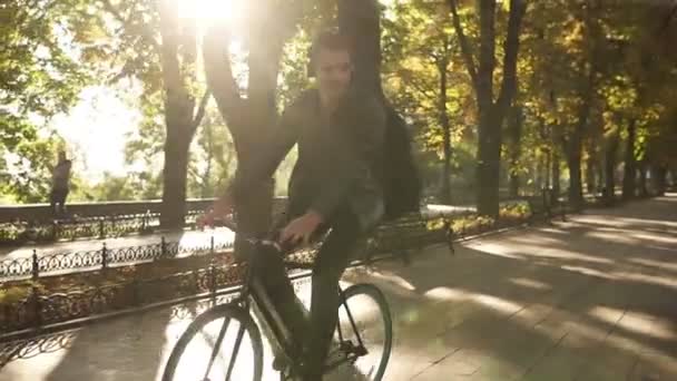 Jovem feliz e sorridente com roupas casuais pretas, fones de ouvido andando de bicicleta pelo parque da cidade pavimentado com guitarra em caixa preta na parte de trás e ouvindo a música. Árvores verdes. Vista frontal — Vídeo de Stock