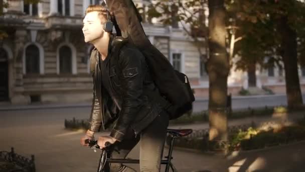 Brunette handsome young man in black casual clothes, headphones riding a bike by the paved city park with guitar in black case on the back and listening to the music. Green trees around. Side view — 图库视频影像