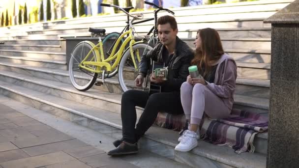 年轻夫妇坐在楼梯上, 男子弹吉他。男子穿着黑色休闲弹吉他, 而与他的女朋友坐在户外的城市楼梯上的格子。他们旁边的自行车。一边 — 图库视频影像