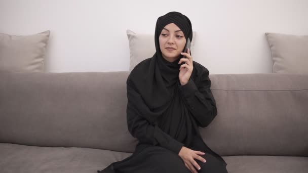 Junge attraktive muslimische Frau in schwarzer Kleidung, die zu Hause auf einer grauen Couch sitzt und lächelnd auf ihrem Handy spricht. Loft-Innenraum. Frontansicht — Stockvideo