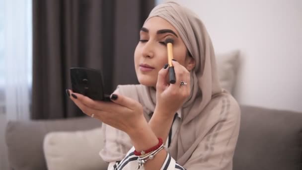 Pandangan samping wanita muslim melakukan makeup di wajahnya dengan kuas, menerapkan eyeshadows. Kehidupan modern Muslim orang, makeup di rumah — Stok Video
