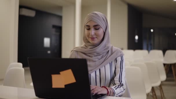 Улыбающаяся уверенная мусульманка в хиджабе, сидящая на белом стуле в современном конференц-зале онлайн с ноутбуком на белом столе — стоковое видео