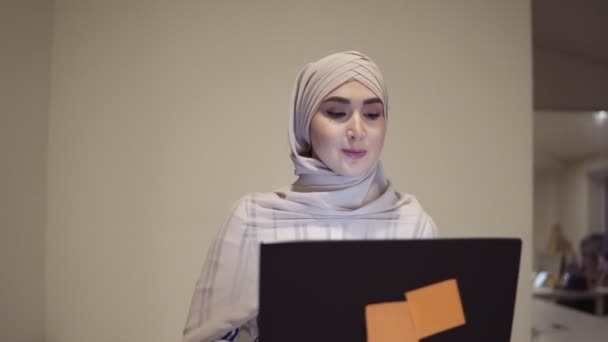 Fiduciosa donna araba che indossa hijab beige andando vicino alla sala mentre tiene il suo computer portatile nero in mano. Impiegato, posto di lavoro, sala conferenze. Vista frontale — Video Stock