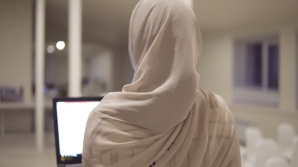 Wanita arab muda mengenakan jilbab di aula sambil memegang laptop hitamnya di tangan. Karyawan, tempat kerja, ruang konferensi, koridor. Tampilan samping, latar belakang yang tidak fokus — Stok Video