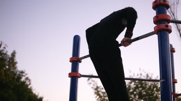 Ein Mann in Sportkleidung turnt draußen auf dem Sportplatz im Park am Reck. Junger Mann macht Klimmzüge in der Natur — Stockvideo