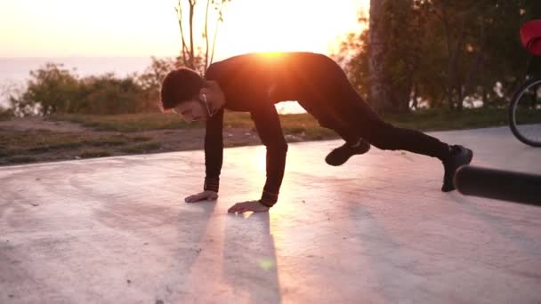 Un joven deportista preparándose para el entrenamiento deportivo y de fitness al aire libre. Concepto de estilo de vida saludable, hombre haciendo flexiones con aplausos. Salida del sol temprano en la mañana en el fondo — Vídeos de Stock