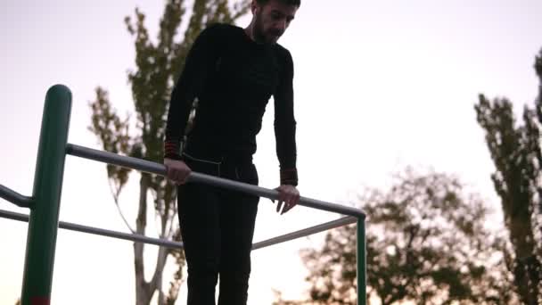 Hombre fuerte, en forma en ropa deportiva negro haciendo ejercicios en la barra horizontal fuera en el campo de deporte en el parque. joven haciendo pull ups fuera de la naturaleza en los auriculares y escuchar a la — Vídeo de stock