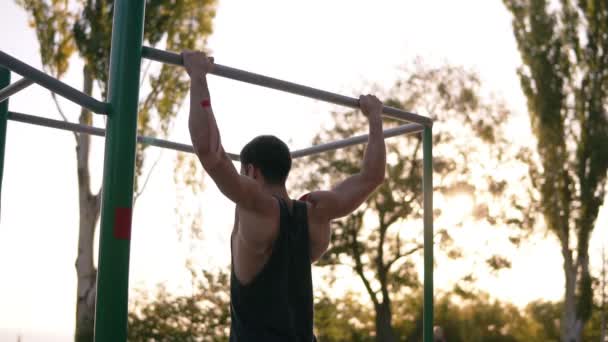 Ajuste o homem muscular na camisa preta fazendo pull-ups no bar horizonte no chão de esportes com árvores e sol brilha no fundo. Movimento lento — Vídeo de Stock