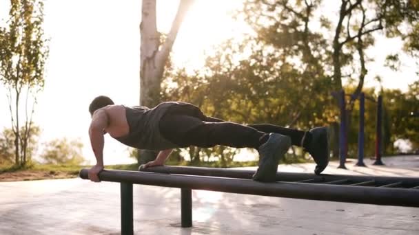 Atletisk ung kaukasiska man göra push ups på barr i lokala utomhus gym i parken. Mannen sport bär svart kläder — Stockvideo