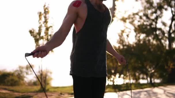 Kaukasischer bärtiger Athlet, der im Freien in schwarzer Sportbekleidung über Springseil springt. Bäume, Natur, Sonnenlicht auf dem Hintergrund — Stockvideo