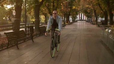 Genç gülümseyen biri sabah park veya boulevard bir bisiklet Bisiklet güneş gözlüğü açık görüntüleri. Ağır çekim Genç adamın bir trekking bisiklet sürme. Yaz, sonbahar boş Şehir Parkı