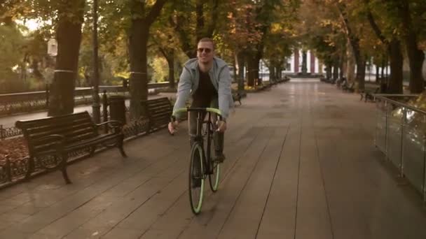 Запись молодого улыбающегося человека в солнечных очках, катающегося на велосипеде в утреннем парке или на бульваре. Медленное движение молодого человека на треккинговом велосипеде. Лето, осень пустой городской парк — стоковое видео