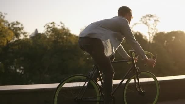 Buon giovane uomo sorridente in occhiali da sole in bicicletta nel parco o viale del mattino. Movimento lento del giovane in sella ad una trekking bike, vento che soffia e agita la camicia. Estate, autunno città vuota — Video Stock