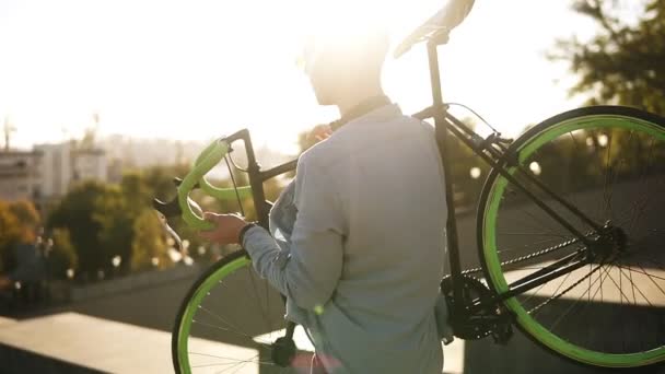 Bonito homem caucasiano jovem em roupas casuais e óculos de sol está segurando uma bicicleta enquanto desce ao ar livre na cidade. Sinalizadores de lente — Vídeo de Stock