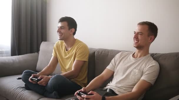 Zwei junge Kerle, die zu Hause Videospiele spielen, Steuerknüppel in der Hand halten und auf dem grauen Sofa im Loft-Innenraum sitzen. — Stockvideo