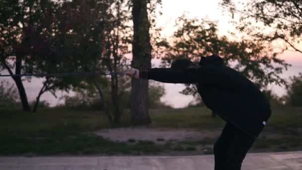 Uomo barbuto in nero sportssuit facendo esercizi di stretching all'aperto per i muscoli della schiena utilizzando banda di resistenza all'aperto in natura. Mattina crepuscolo, sagome di alberi sullo sfondo. Vista laterale — Video Stock