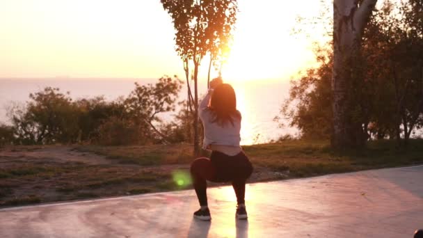 Active woman exercising and stretching in city park in front the sea or ocean. Attractive fit girl training squatting at morning outdoors. Healthy, fitness, wellness lifestyle. Rare view — Stock Video