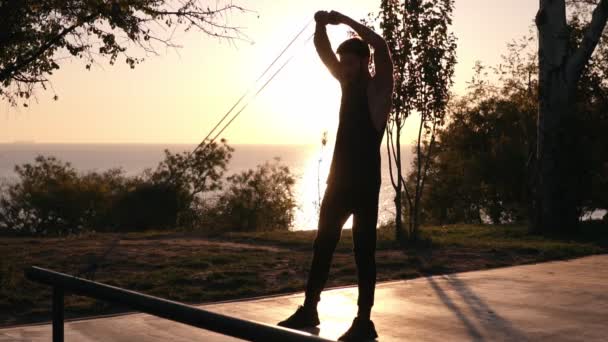 Silhouette d'un homme faisant des exercices de bras et squats à l'extérieur en utilisant une bande de résistance à l'extérieur dans la nature. Triceps et jambes exercices avec du caoutchouc. Lever de soleil matinal. Mouvement lent — Video