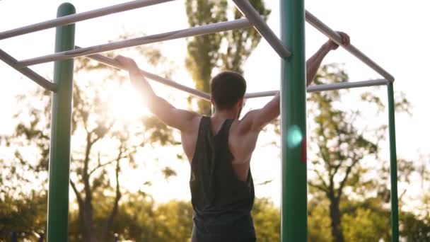 Hombre musculoso fuerte en camisa negra haciendo difíciles pull-ups en la barra del horizonte en el campo de deportes con árboles y el sol brilla en el fondo. Movimiento lento — Vídeo de stock