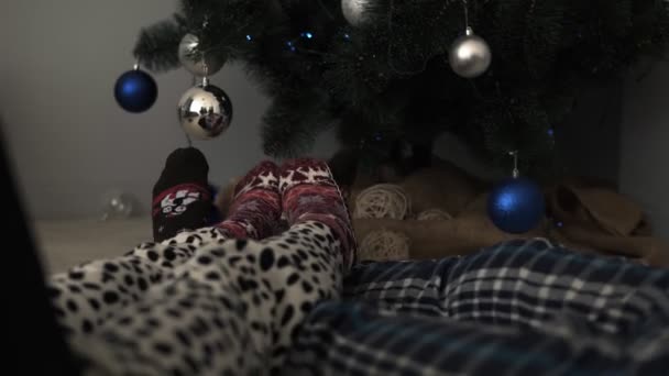 Detalle de los pies masculinos y femeninos con calcetines cálidos de invierno, colocados en el suelo con un árbol de Navidad bien decorado y juguetes de Navidad en el fondo. Divertirse juntos — Vídeos de Stock