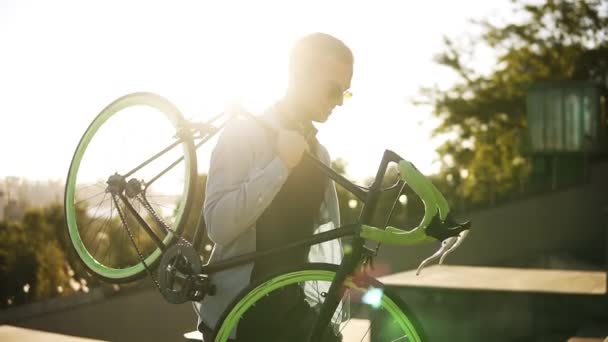 Guapo joven caucásico en ropa casual y gafas de sol está sosteniendo una bicicleta un hombro mientras sube al aire libre en la ciudad. Lente destellos en el fondo — Vídeos de Stock