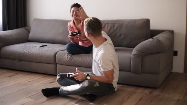 Pareja joven jugando videojuegos en casa, sentado en el sofá y el hombre en el suelo, disfrutando de tiempo juntos jugando con joysticks. El hombre ganó el juego y le dio cinco a su novia. — Vídeos de Stock