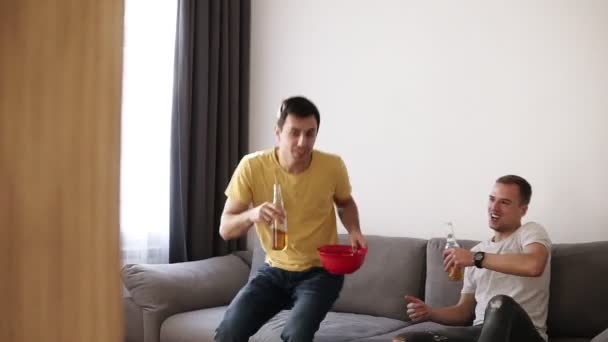 Foto de dos amigos que vienen a la sala de estar viendo la televisión sentados en un cómodo sofá en casa, comiendo papas fritas del tazón y bebiendo cerveza — Vídeo de stock