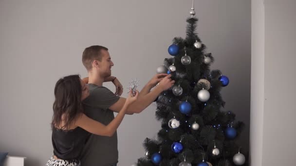 Couple jeune mari et femme se préparant pour le Nouvel An ou Noël. Embrasser debout près du sapin et décorer avec de l'argent et des jouets bleus puis embrasser. À l'intérieur — Video