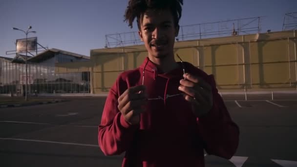 Elegante hombre joven de piel oscura con tonos de moda y camisa roja sosteniendo luego se puso las gafas de sol. De pie fuera en la zona de aparcamiento — Vídeos de Stock