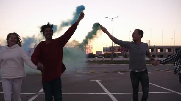 Vista frontal de cuatro locos amigos jóvenes de raza mixta corriendo con coloridas bombas de humo en las manos en la zona de estacionamiento vacía afuera. Emociones, estilo de vida feliz — Vídeos de Stock