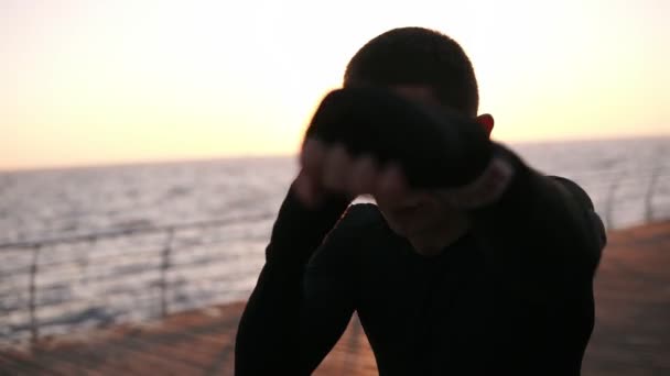 Vue de face d'un boxeur masculin en chemise noire pendant le processus d'entraînement sur la promenade en face de la mer tôt le matin. Portrait d'un homme boxant avec un adversaire invisible, frappant - portant du blanc — Video