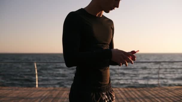 Hombre atlético joven en negro T interruptor en su pista favorita en su teléfono inteligente. Escuche música frente al mar al aire libre en verano y comience a correr. Calentamiento — Vídeo de stock