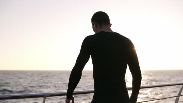 O jovem corredor parou para descansar um pouco antes da corrida de longa distância. Ele usa uma camisa preta esportiva com fones de ouvido brancos. De manhã cedo, em frente ao mar. Vista rara — Vídeo de Stock