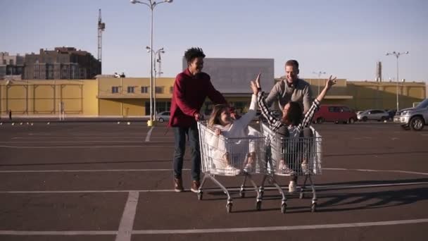 Vista frontal de un joven amigo divirtiéndose al aire libre en carritos de compras. Jóvenes multiétnicos corriendo en carritos de compras en la zona de estacionamiento con sus novias. Pareja riendo, teniendo tiempo — Vídeo de stock