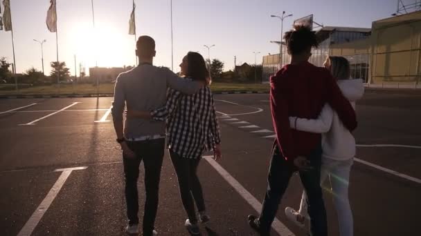 Groep van jonge multirace paren lachen tijdens het wandelen door de asfaltweg. Jonge mannen en vrouwen die genieten van een zomerdag op parkeerzone in de buurt van het winkelcentrum. Vooraanzicht, zon schijnt op de achtergrond. Zeldzame weergave — Stockvideo